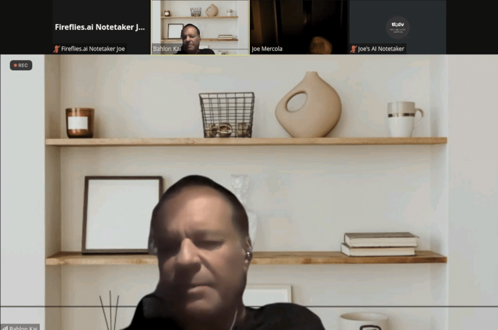 A screen shot from a video. Kai Clay has his eyes closed. He's on a zoom call with other attendees visible across the top and an artificial background behind him. 